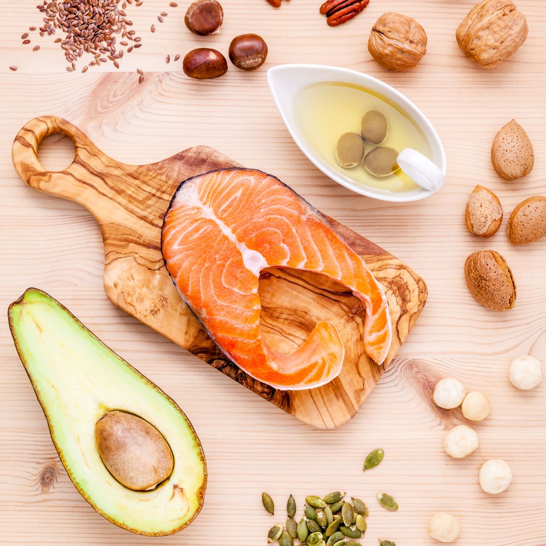 Sélection d'aliments riches en acides gras sains, comprenant du saumon, des noix, des graines, de l'huile d'olive et un avocat sur une planche en bois.